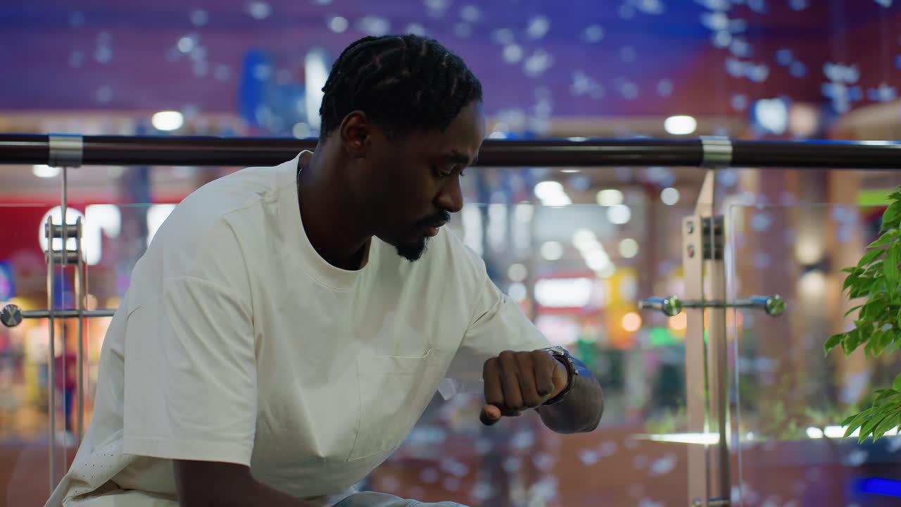 Man in white shirt and jeans seated on bench inside shopping mall holding hands together with anxious expression while waiting for someone and checking time near illuminated green plant modern retail