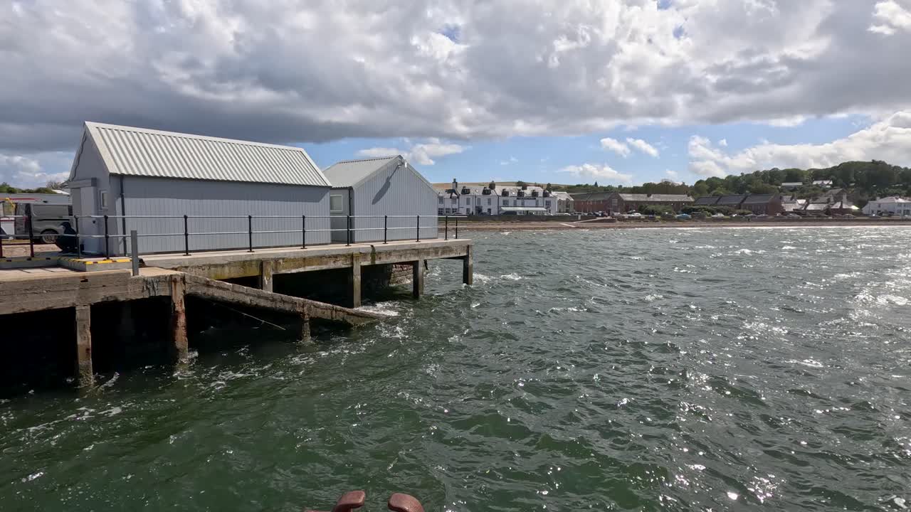 Waves ripple across greenish sea as the camera pans past a pier and metal waterfront building under dramatic, partly cloudy skies in Cromarty, Scotland