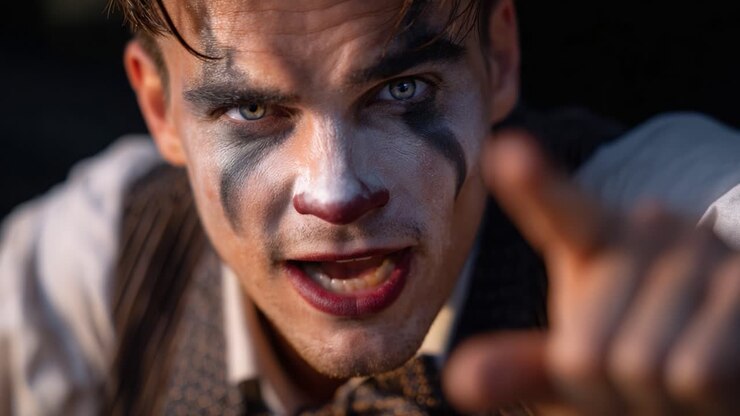 Primo piano intenso di un personaggio simile a un clown che fa un cenno con un'espressione severa, mettendo in mostra un trucco vivace e uno sguardo accattivante, evocando fascino e inquietudine