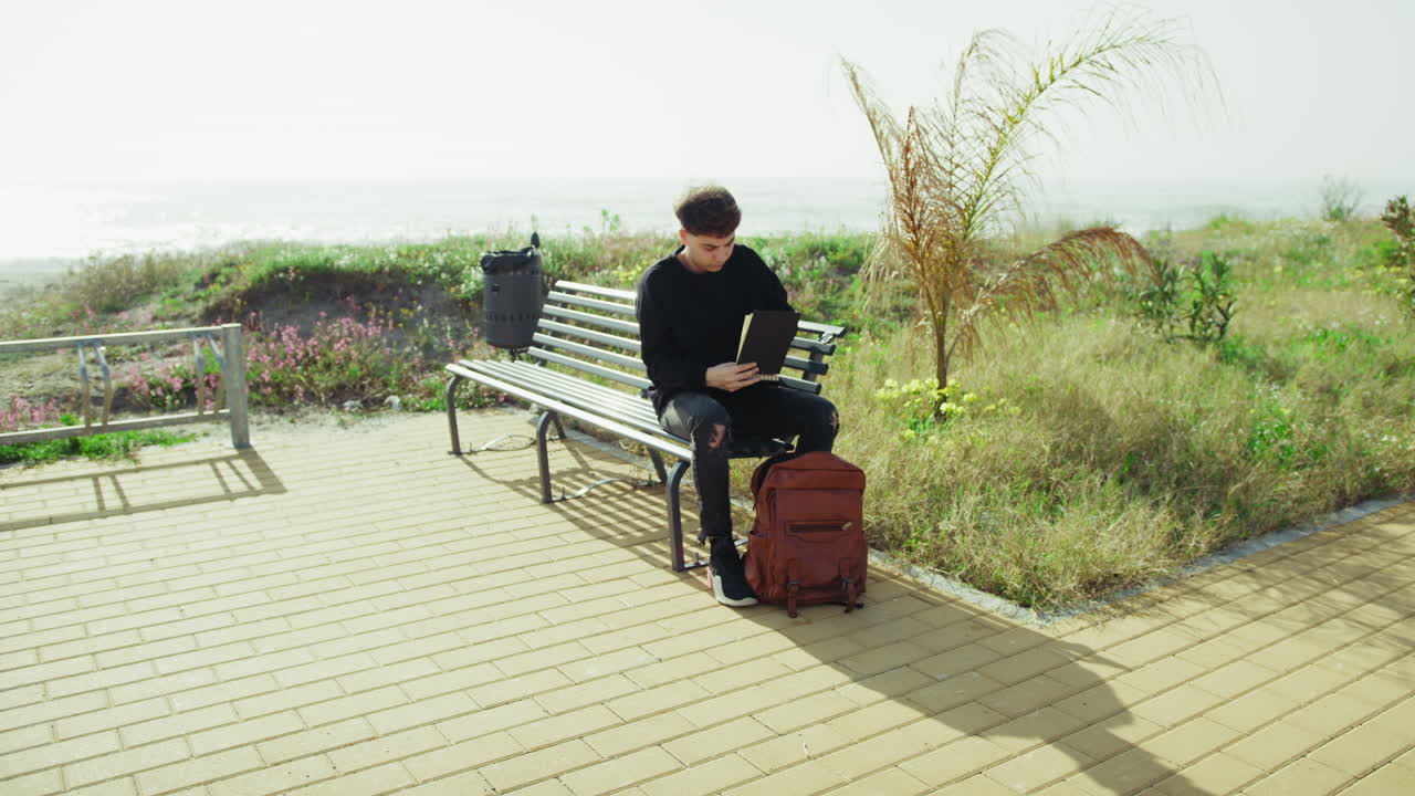 Young Man On A Bench Near The Sea Picking A Notebook From The Backpack
