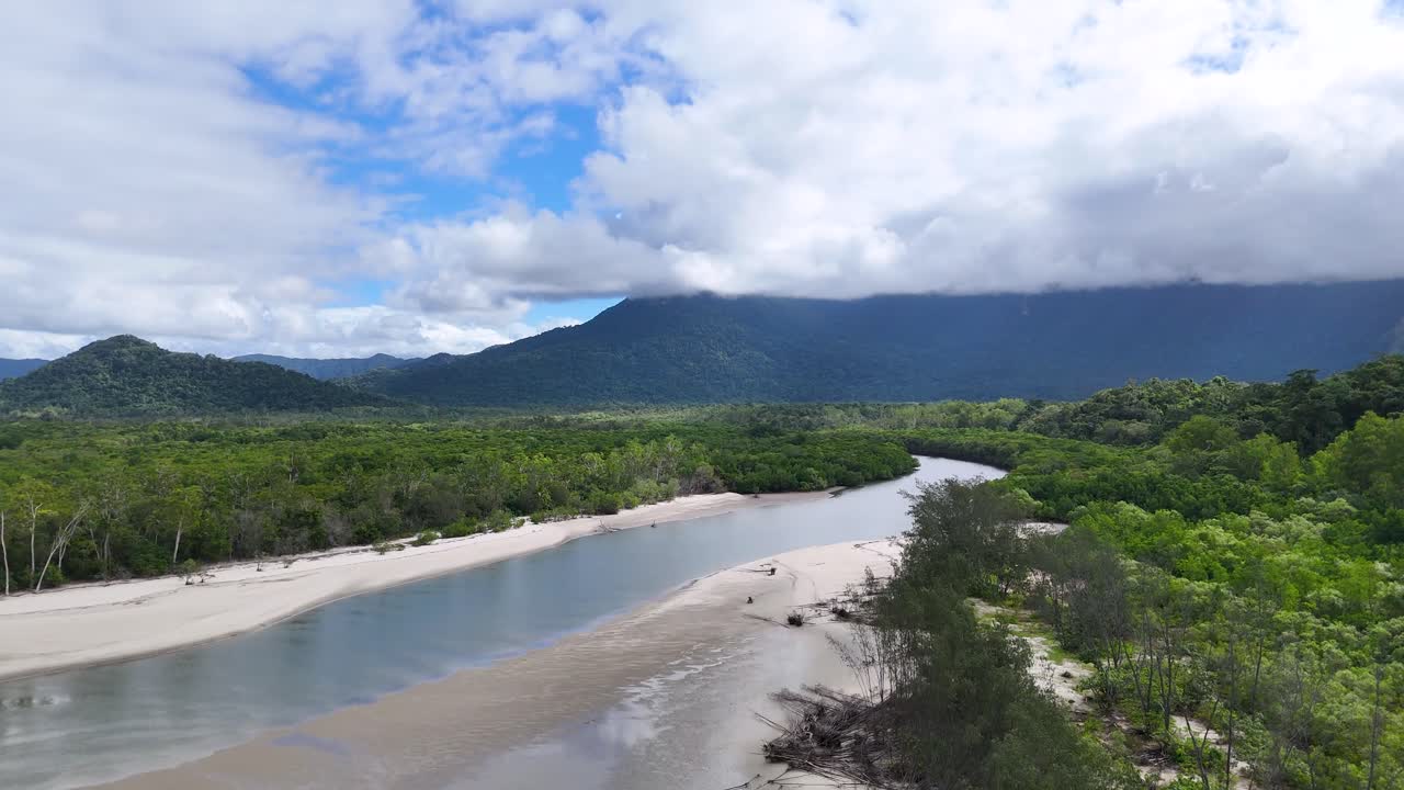 Drone pans above sandy river bend, lush rainforest, and distant mountains under bright daylight