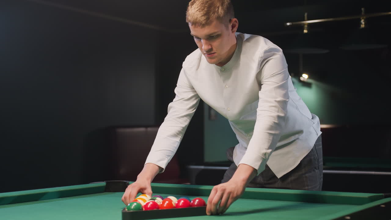 Young man in white shirt and gray trousers carefully arranges colorful billiard balls in triangle formation on vibrant green pool table, preparing for precise gameplay in a modern billiard hall