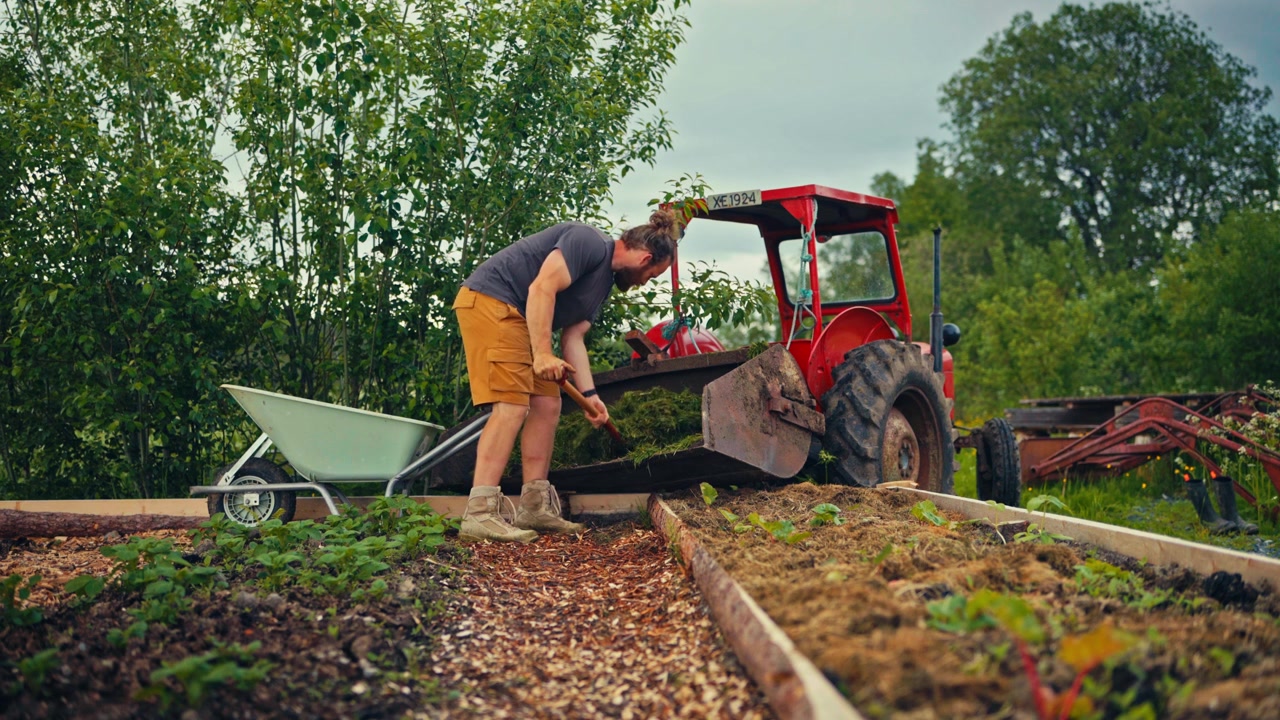 A man works beside a tractor on a rural farm in Norway, appearing to prepare garden beds or fertilize fields in an agricultural setting