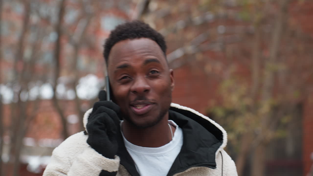 Homme conversant joyeusement au téléphone, souriant, avec des arbres dénudés et un bâtiment en arrière-plan, dans un cadre extérieur décontracté en hiver, exprimant sa joie en conversant.