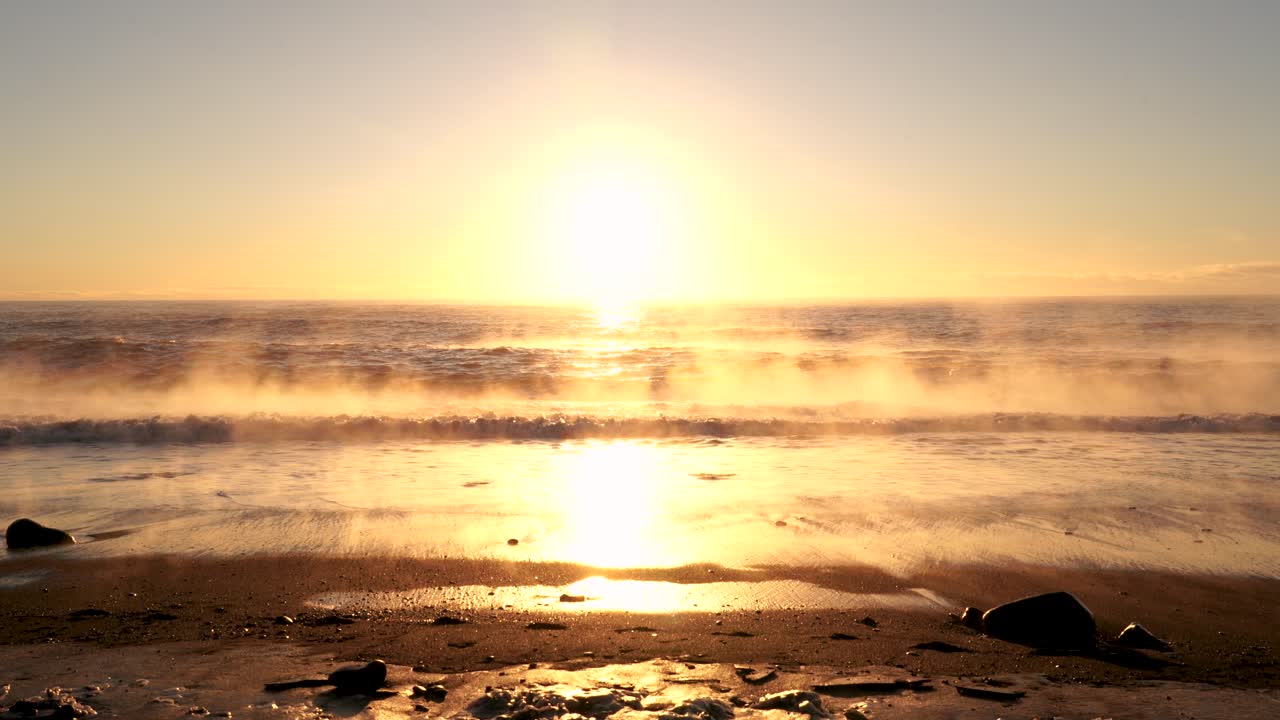 신비한 안개와 함께 황금빛 일몰 동안 검은 해변에 도달하는 파도의 넓은 샷
