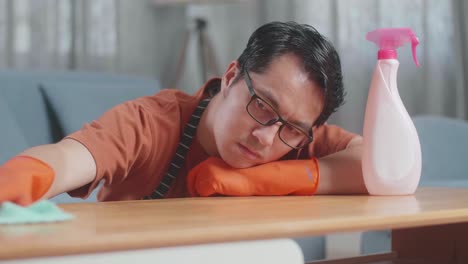close up of bored asian male housekeeper with an apron cleaning the table by the spray at home