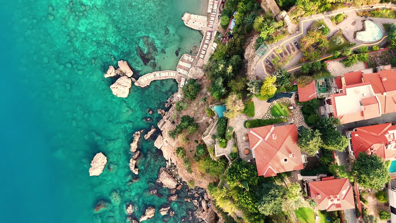 Stunning bird's eye view of the turquoise Mediterranean coastline, featuring rocky beaches, cliffside terracotta-roofed buildings, and crystal-clear waters.
