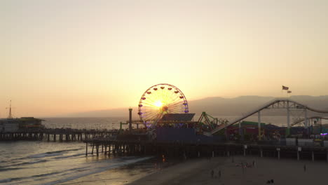 Santa-Monica-Pier-Bei-Wunderschönem-Sonnenuntergang-Mit-Amerikanischer-Flagge-Und-Kalifornischer-Flagge,-Die-Am-Riesenrad-Des-Themenparks-Mit-Meerblick,-Weitem-Luftblick,-Dollyrutsche-Rechts-Weht