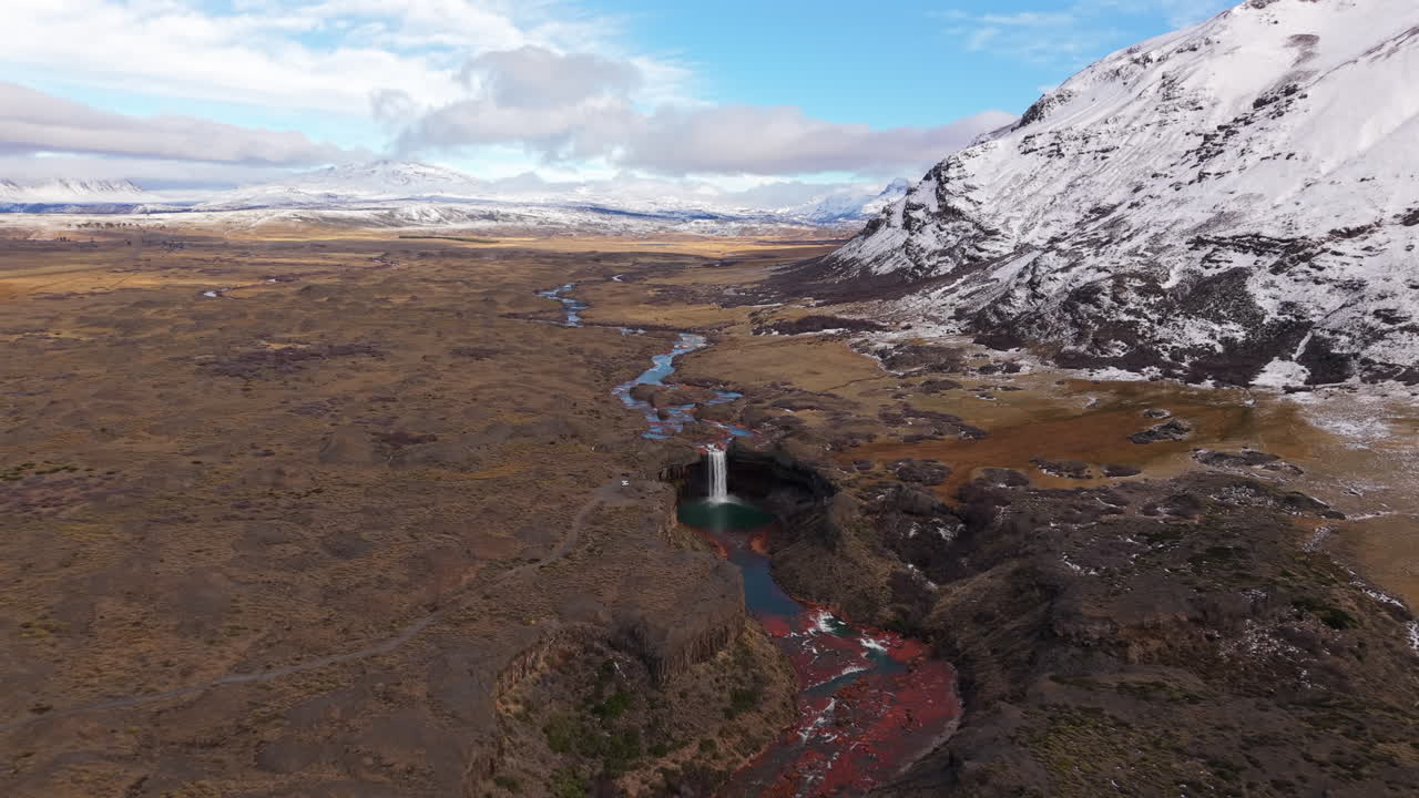 Drone pull away reveals Salto del Agrio in Caviahue, a 60m waterfall plunging into a green pool, framed by vivid basalt cliffs and ancient araucaria trees on the Patagonian Andes plateau