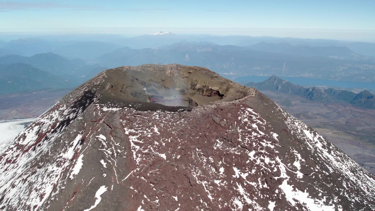 En hisnande utsikt från toppen av en vulkan avslöjar en stor krater omgiven av stenig terräng, med en fridfull sjö synlig i fjärran mot en klarblå himmel