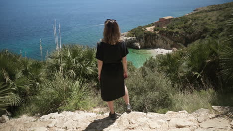 woman stands on cliff by turquoise blue beach in sicily, italy