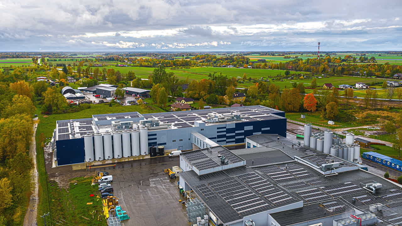 Drone hyperlapse over a large factory with rooftop solar panels and storage silos beside autumn farmland under cloudy sky, showing sustainable manufacturing and rural industry landscape