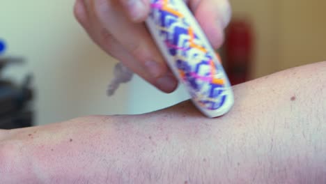 pequeña maquinilla de afeitar eléctrica de mano blanca con patrón de diseño púrpura y naranja, afeitando el antebrazo de un hombre o mujer caucásicos blancos con pecas en el brazo