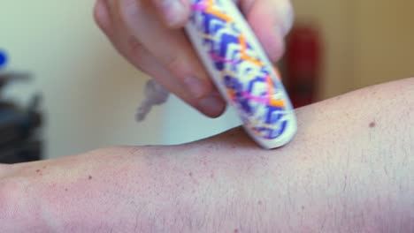 little handheld electric razor white with purple and orange design pattern, shaving the forearm of a white caucasian man or women with freckles on arm