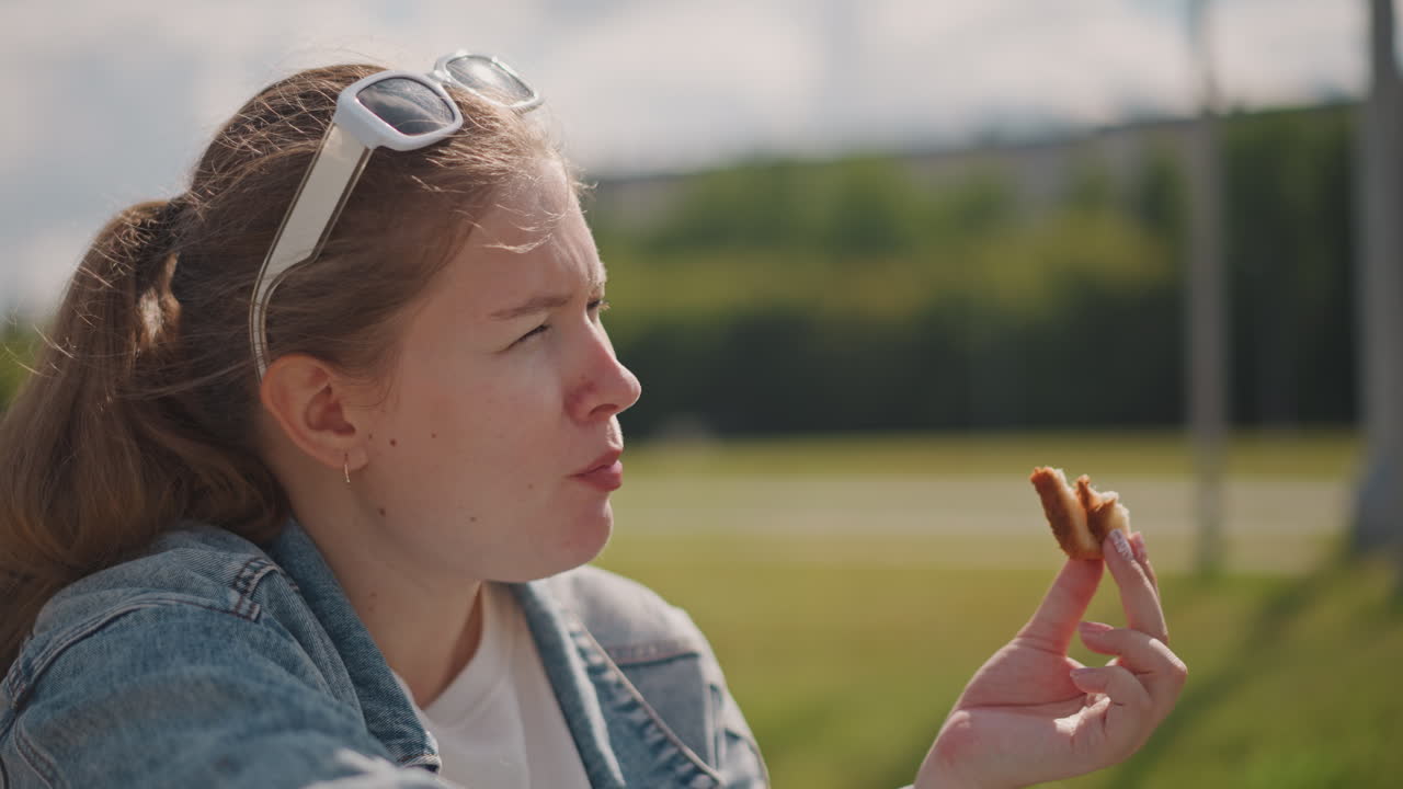 Primo piano di donna caucasica che assaggia un dolce, sguardo contemplativo verso l'orizzonte, briciole sulle labbra, occhiali da sole sulla testa, luce solare delicata sulla pelle, sfondo tranquillo di un parco, respiro sottile e pensierosa.