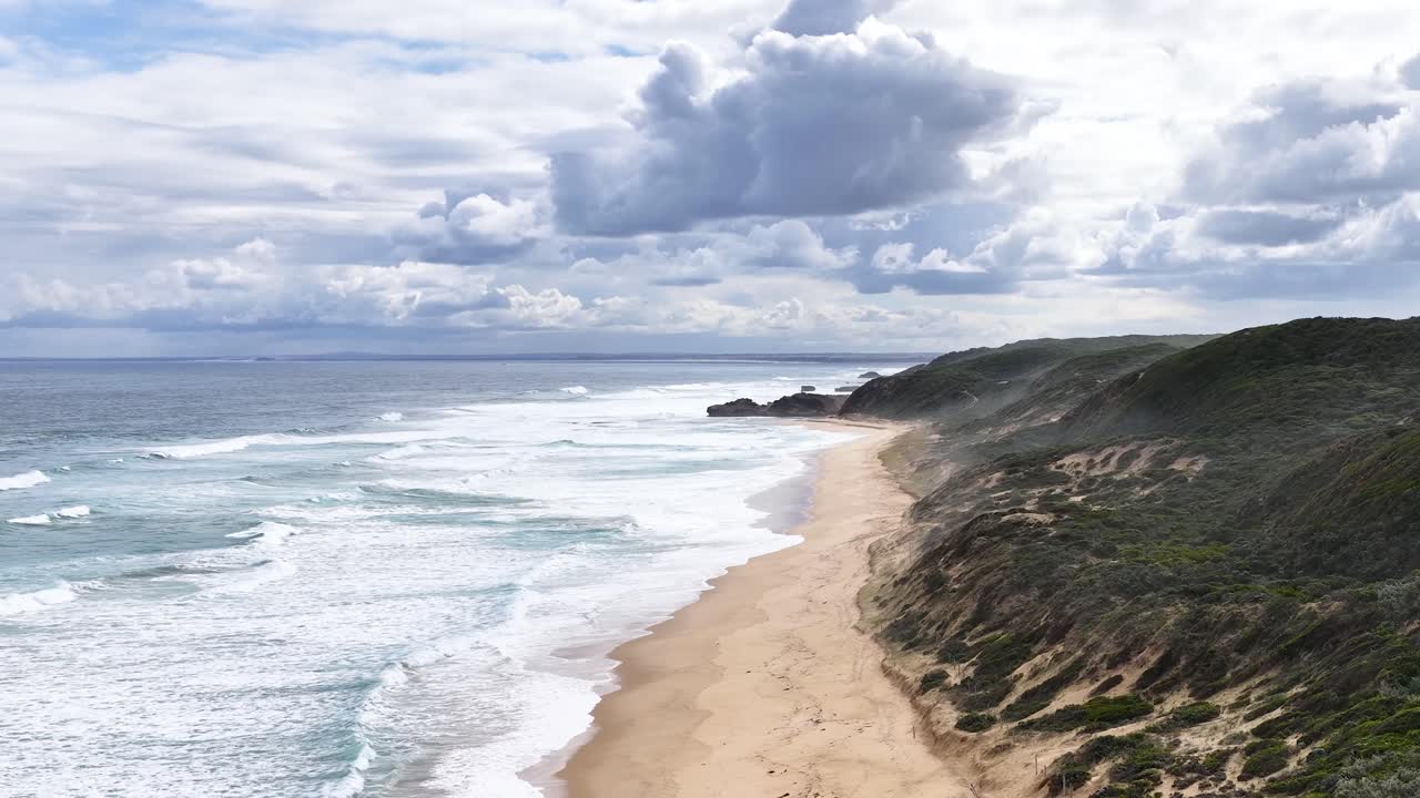 Drone camera glides above sandy beach, capturing waves, rugged cliffs, and dramatic cloudy sky