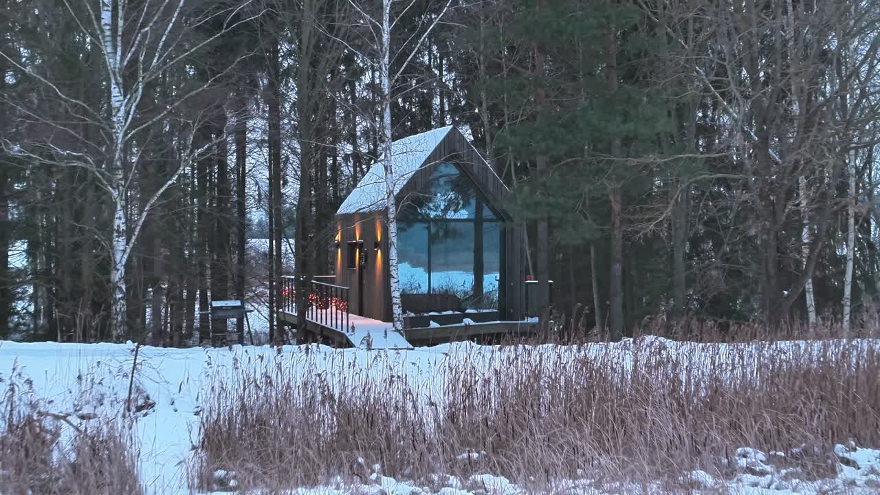 Ripresa con drone che si muove verso una moderna baita di vetro circondata da una foresta innevata a Tervete, Lettonia. Architettura minimalista fusa con un ambiente boschivo invernale