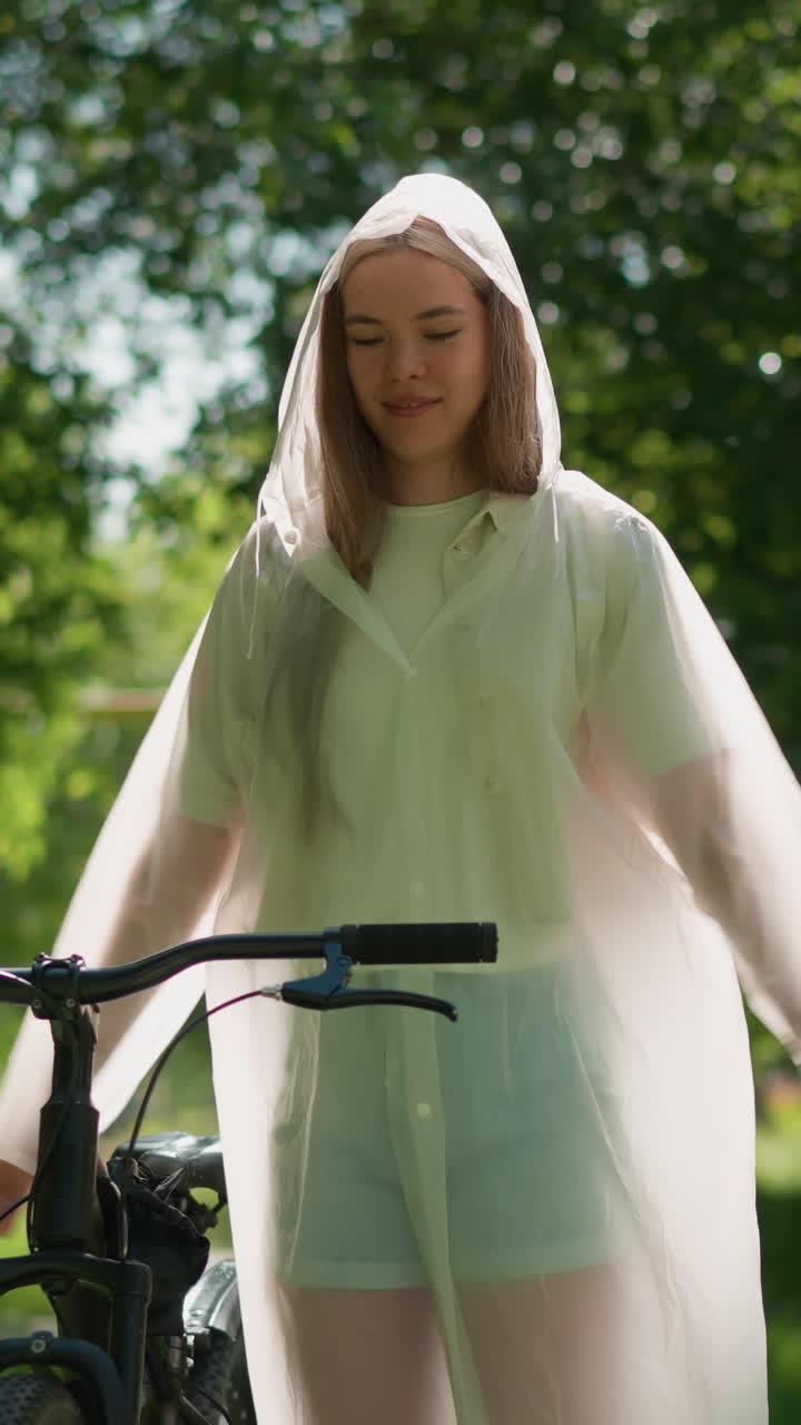 Young woman in translucent raincoat stands near bicycle with arms outstretched, joyfully swinging her hands as if wanting to fly, background filled with lush greenery