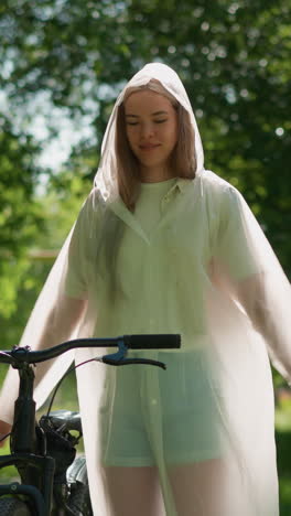 mujer joven en impermeable translúcido se encuentra cerca de la bicicleta con los brazos extendidos, alegremente balanceando sus manos como si quisiera volar, el fondo lleno de vegetación exuberante