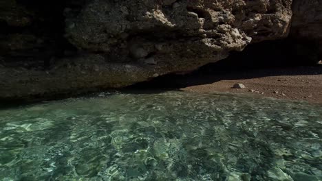 Crystal-sea-water-splashing-under-cliffs,-reflecting-sunlight-on-seabed-with-white-pebble,-beautiful-bay-in-Mediterranean