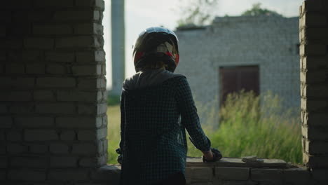 back view of a lady standing near a window of an old building, resting her hand on the ledge, and looking around towards an abandoned house overgrown with grass in a rural area