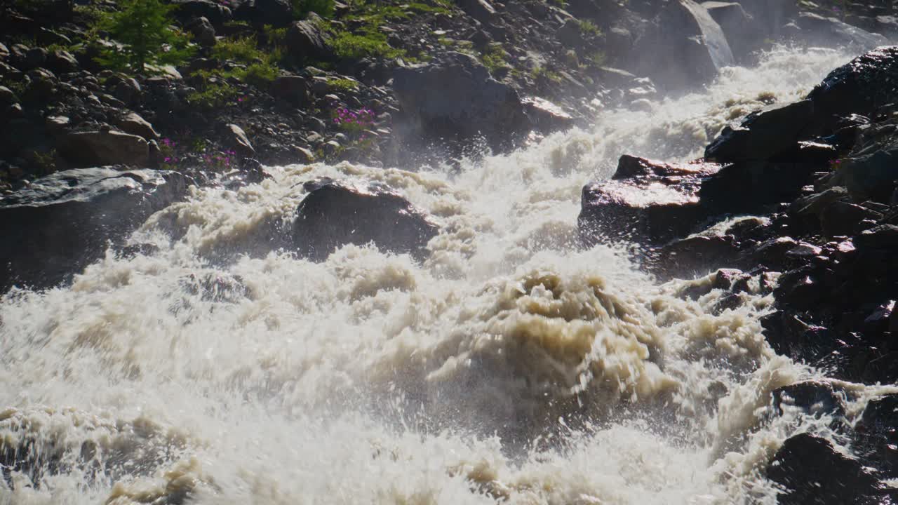 Une rivière puissante se précipite sur des rochers sombres. L'eau s'écrase, projetant de la brume dans l'air. La lumière du soleil se reflète sur la surface agitée. La scène capture l'énergie d'une nature sauvage et indomptée. Idéal pour l'aventure, les activités de plein air.
