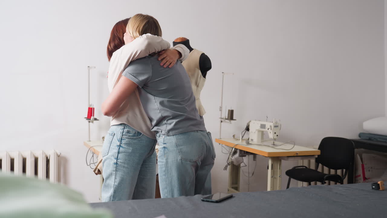 Seamstress duo hug inside sewing studio, sharing triumphant high five beside industrial machines, celebrating project success with lively body language and denim clad legs in creative workspace