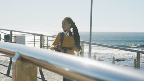 Afroamerikanische-Frau-Mit-Kopfhörern-Und-Rucksack,-Die-Auf-Der-Promenade-Am-Meer-Roller-Fährt