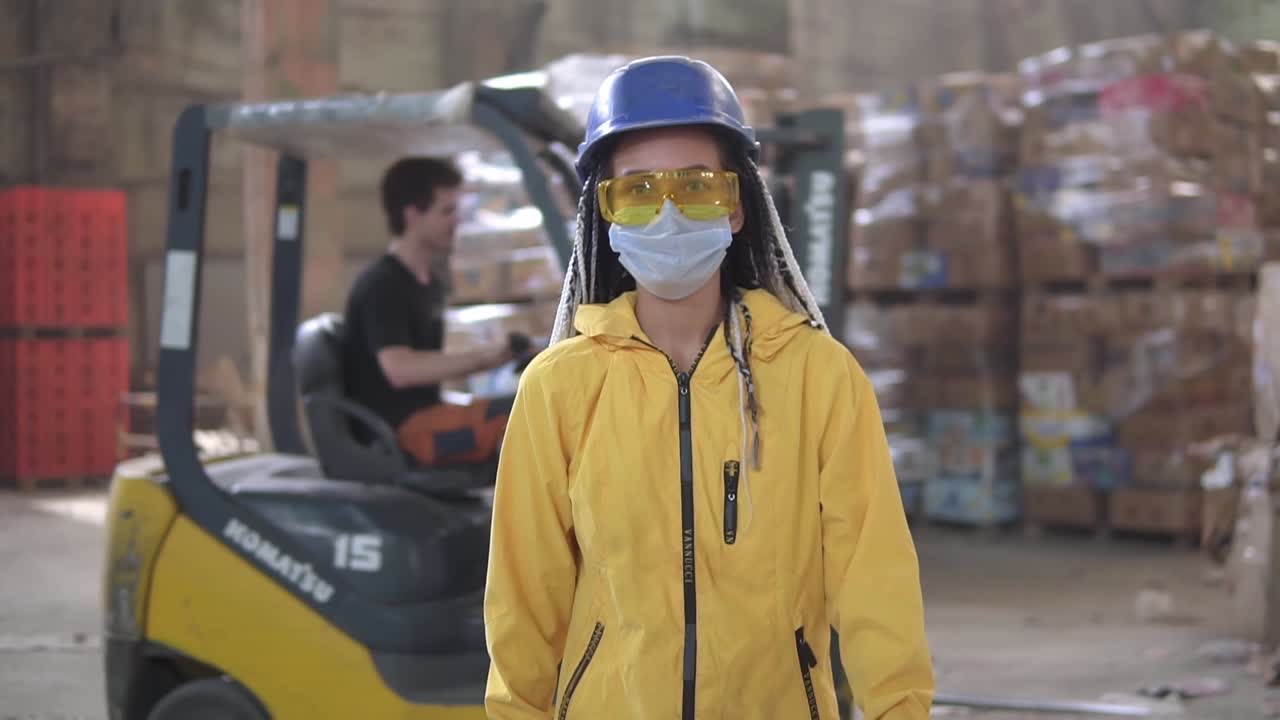 폐기물 처리 공장. 추가 폐기물을 재활용하고 저장하는 것. 강한 모자, 장갑과 마스크를 입은 여성 노동자가 압축 폐기물  ⁇ 어리, 거대한  ⁇ 어리를  ⁇ 고  ⁇ 고 있습니다. 앞면 영상. 배경에 로딩 기계