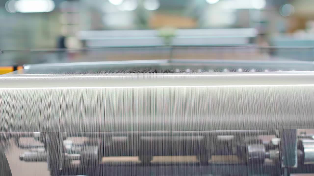 A close-up shot shows a high-speed industrial loom automatically weaving thousands of fine white threads into fabric at a large textile manufacturing plant