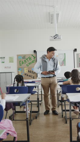 vertical video of happy diverse male teacher and schoolchildren writing in classroom in slow motion
