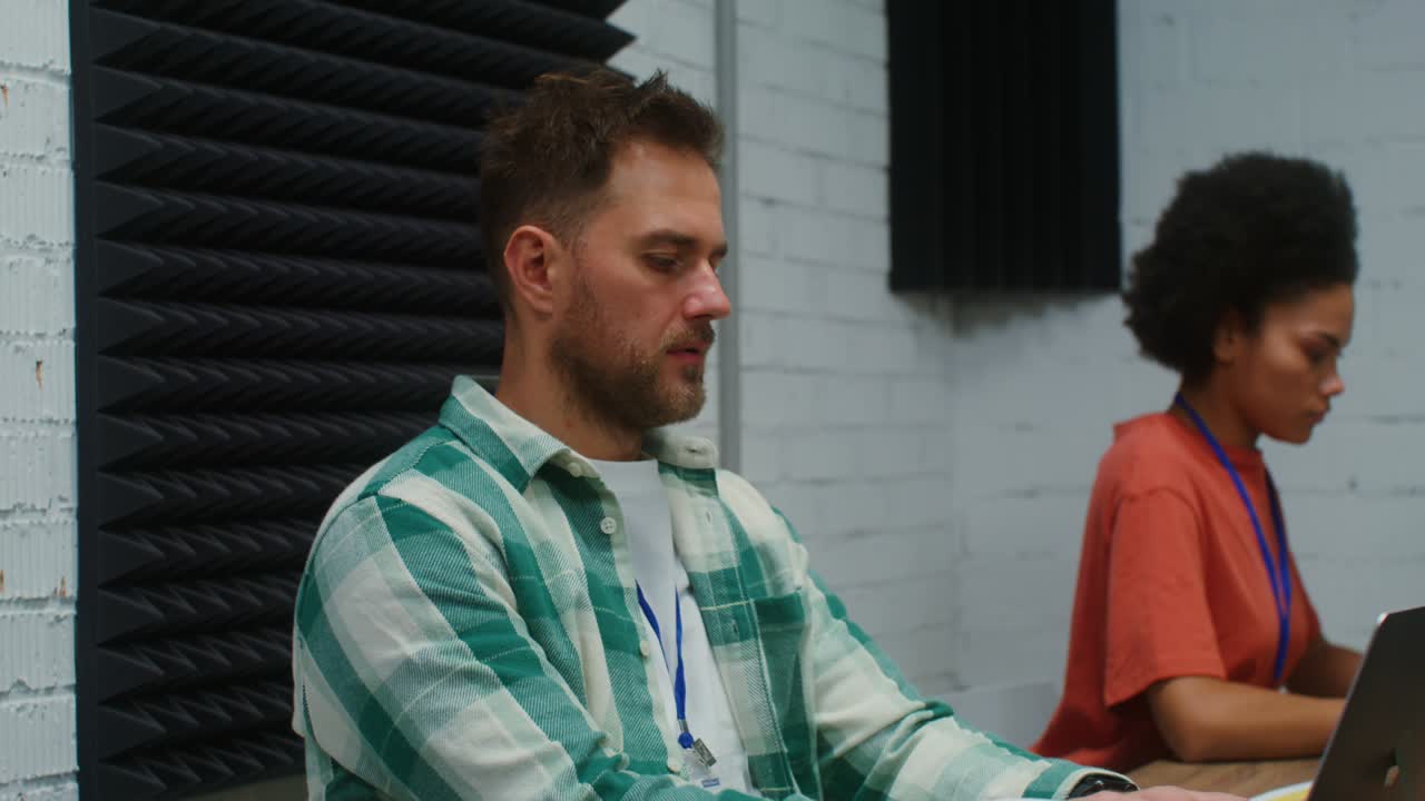 A man and woman work at laptops, sitting at one work desk in a office