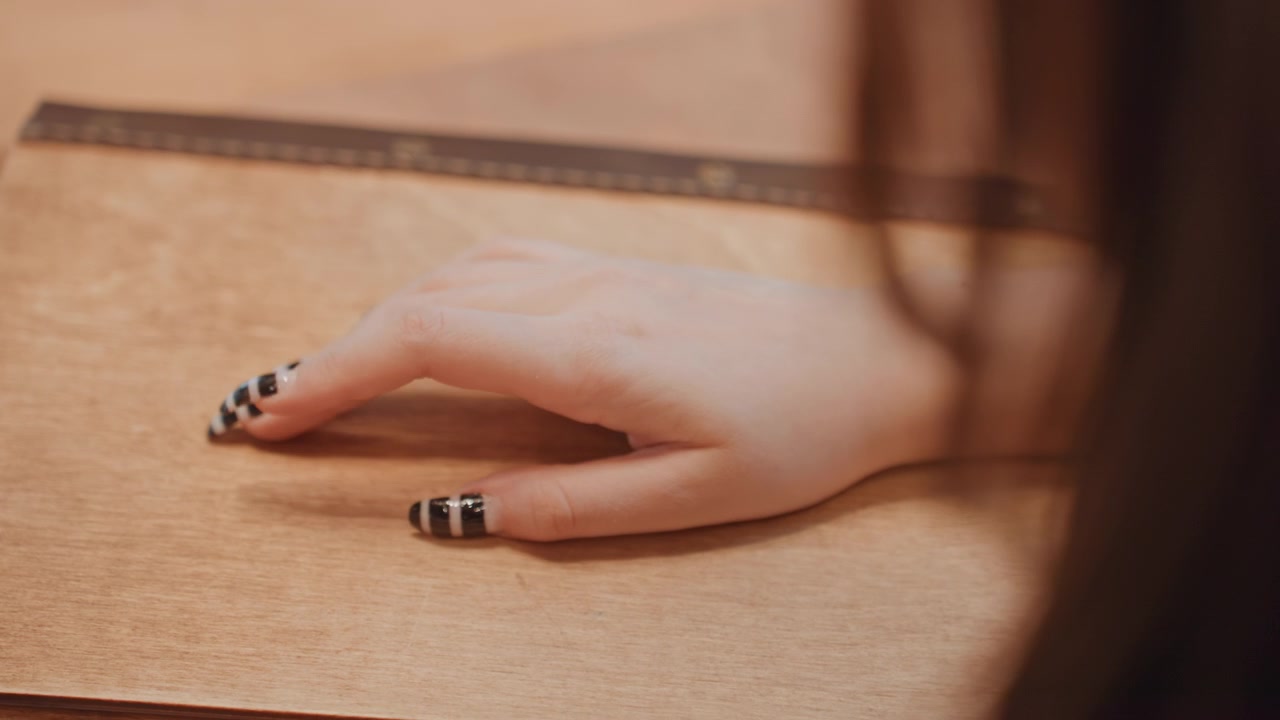 Mano di scrittrice su un diario di pelle, primo piano di unghie curate con smalto a strisce nere, righello in evidenza, scrivania in legno caldo, illuminazione ambientale soffusa, umore contemplativo, gesto di pianificazione creativa.
