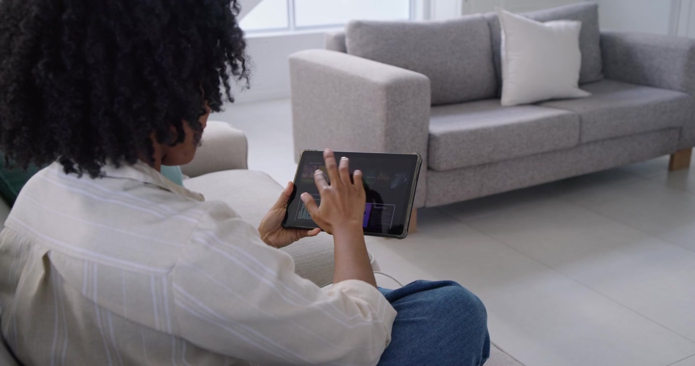 Femme afro-américaine inclinant une tablette, tapant et faisant glisser pour choisir du contenu sur un canapé gris