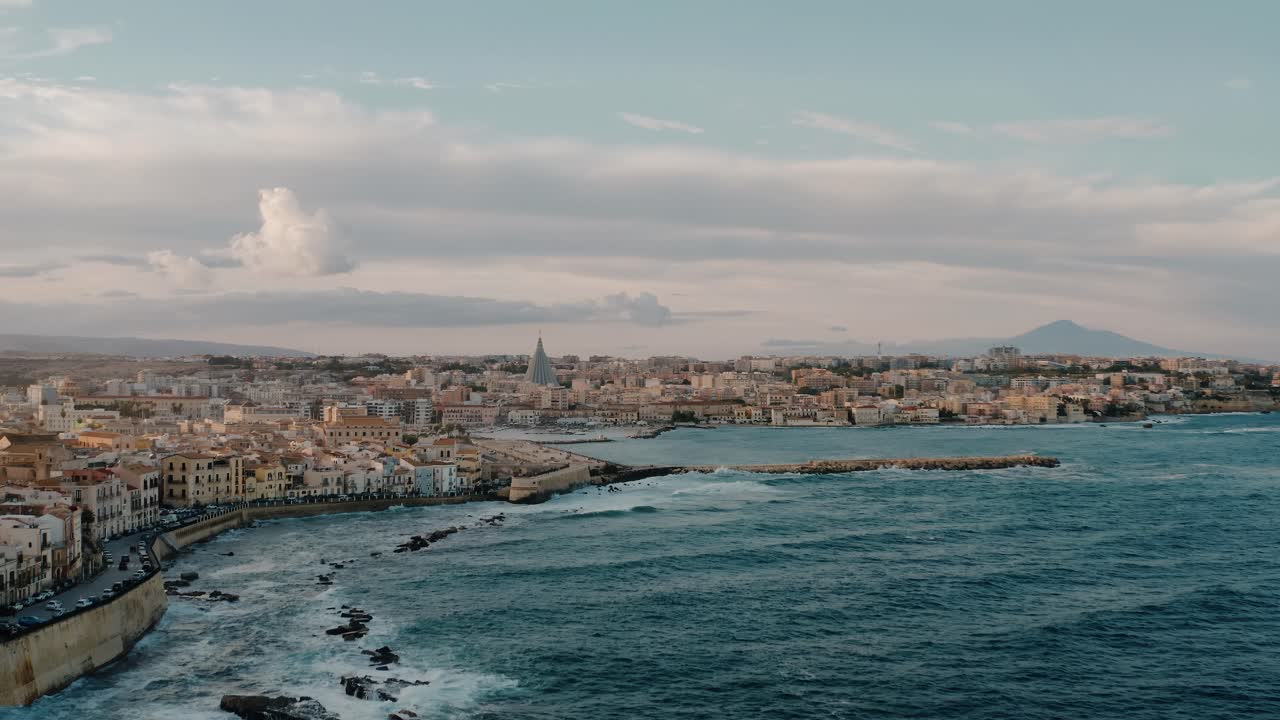 Wide drone shot of Ortigia island and Mount Etna in a distance. Aerial view over historic buildings on a natural fortress by Mediterranean sea with waves crashing on the coast. UNESCO. Sicily. Italy.