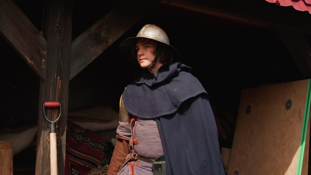 Rievocatore in costume con elmo e mantello scuro si muove attraverso un rifugio di legno in ombra, tiene un sacco pesante sull'uscio, poi prosegue attraverso il cortile di un villaggio rustico sotto una luce soffusa e nuvolosa.