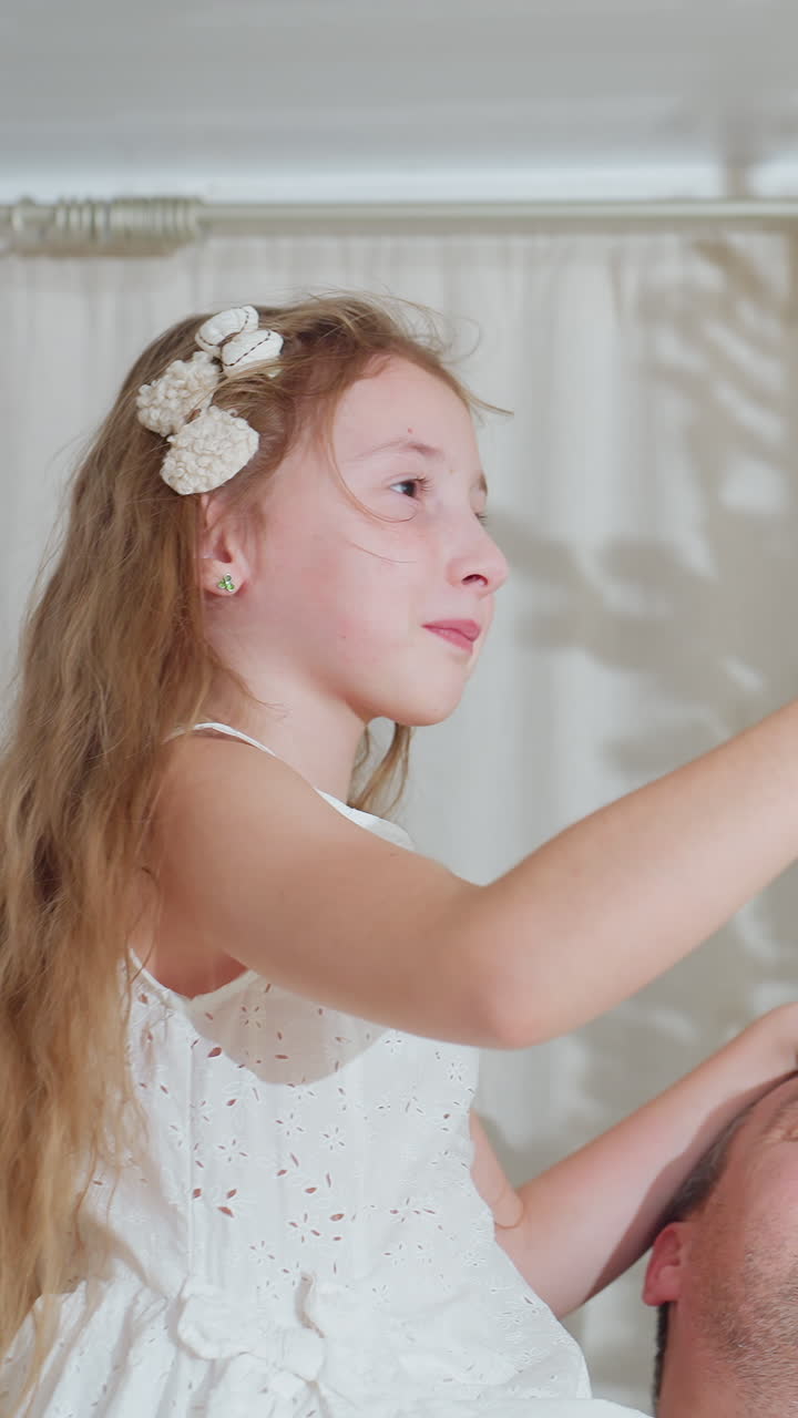 Niña caucásica sobre los hombros de su padre alcanzando un adorno en el interior de su casa, con un vestido blanco calado y un clip floral en el pelo, largos rizos castaños, expresión alegre, apoyo afectuoso de su padre, suave