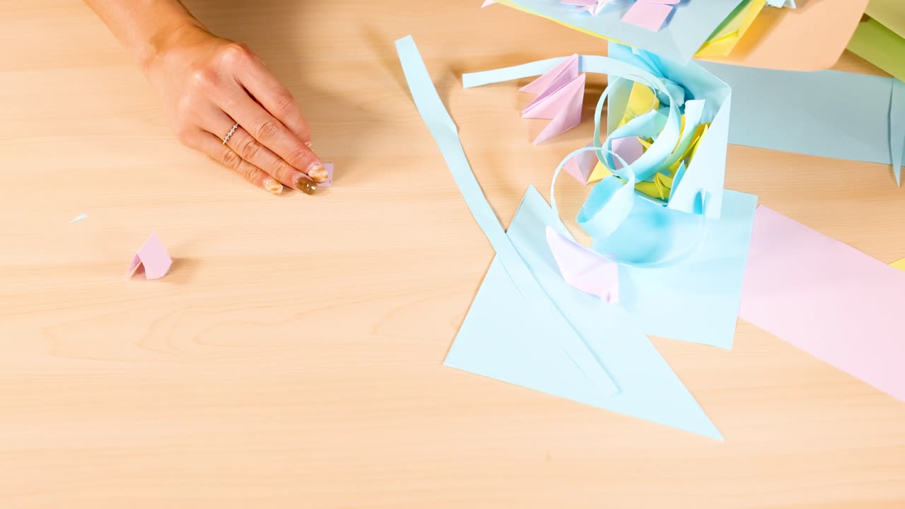 Person folds pastel origami paper on wooden table, overhead view, bright even lighting, static camera