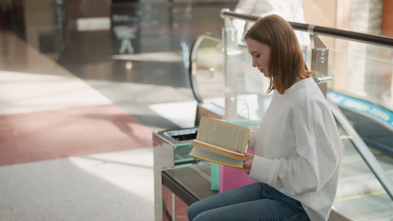 vista lateral de una mujer sentada en un centro comercial leyendo un libro cubierto de amarillo con escaleras mecánicas en movimiento y una figura borrosa pisando la escalera mecánica en el fondo