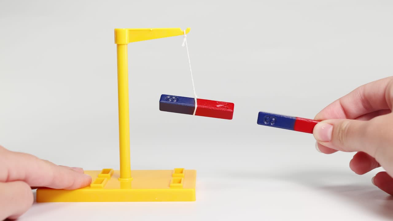 Hands manipulate magnets to show attraction and repulsion, using a suspended magnet setup. Bright lighting highlights the magnetic forces