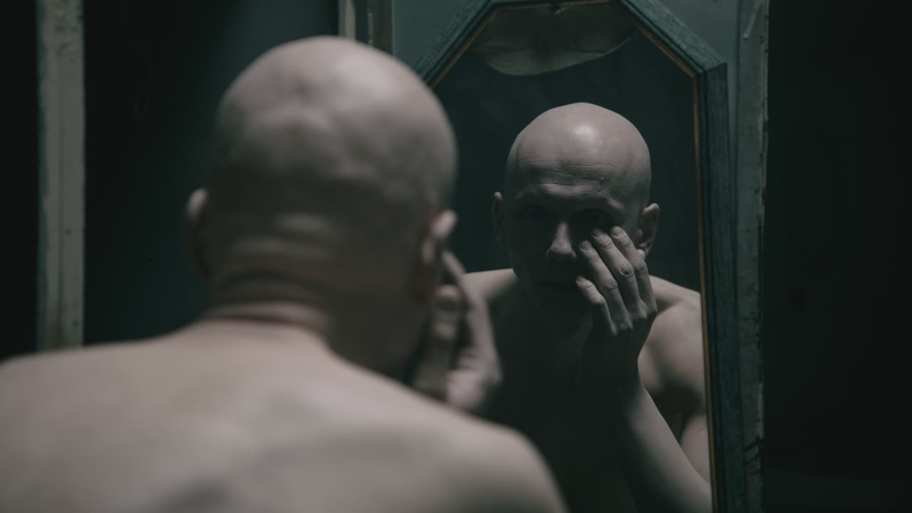 Pale bald skinny man looks into the mirror in the creepy old bathroom at night