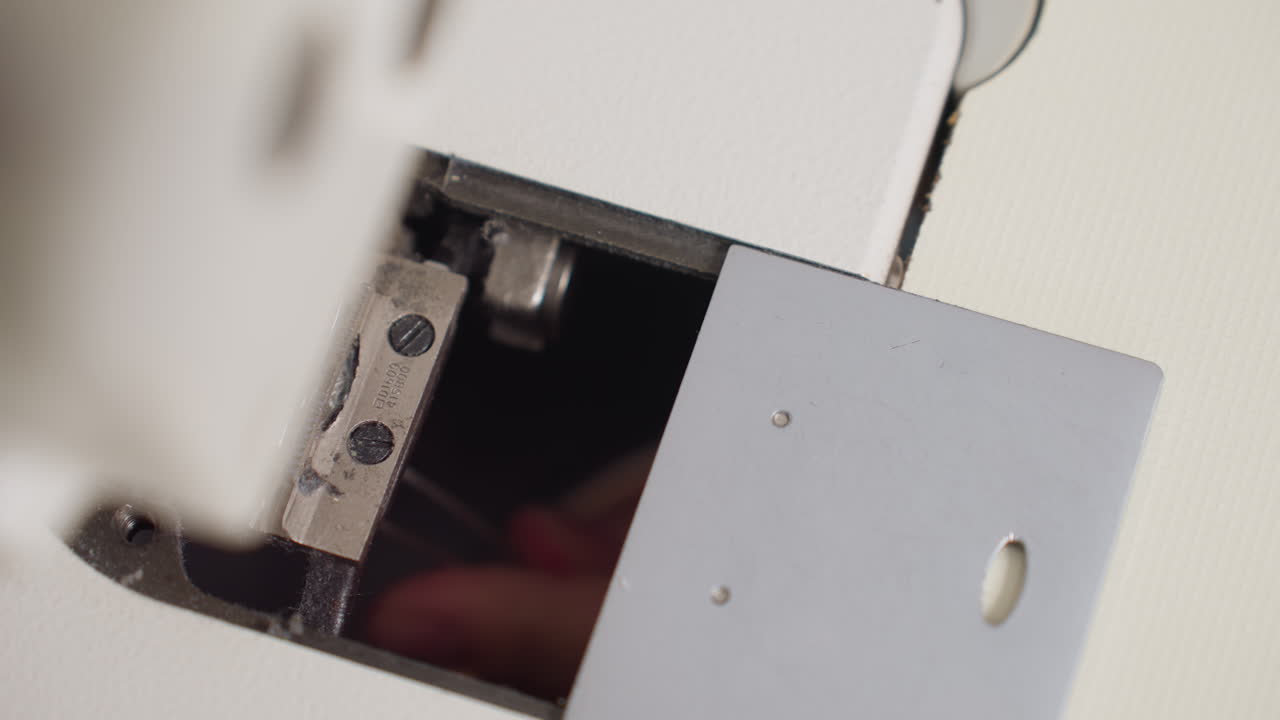 Close up under sewing machine showing hand clipping excess thread from feed dog area, highlighting precision work, mechanical detail, and maintenance care in bright workspace with soft surface
