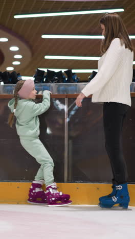 una vista cercana de una niña joven y su madre de pie en una pista de hielo, la niña está vestida con un traje verde menta y patines rosados, detrás de ellos hay una fila de patines de alquiler y un fondo festivo borroso