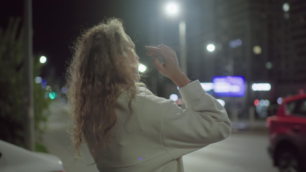 Woman in white coat stands by roadside under streetlights at night adjusting hair while car drives past on urban street with illuminated background signs and blurred city lights
