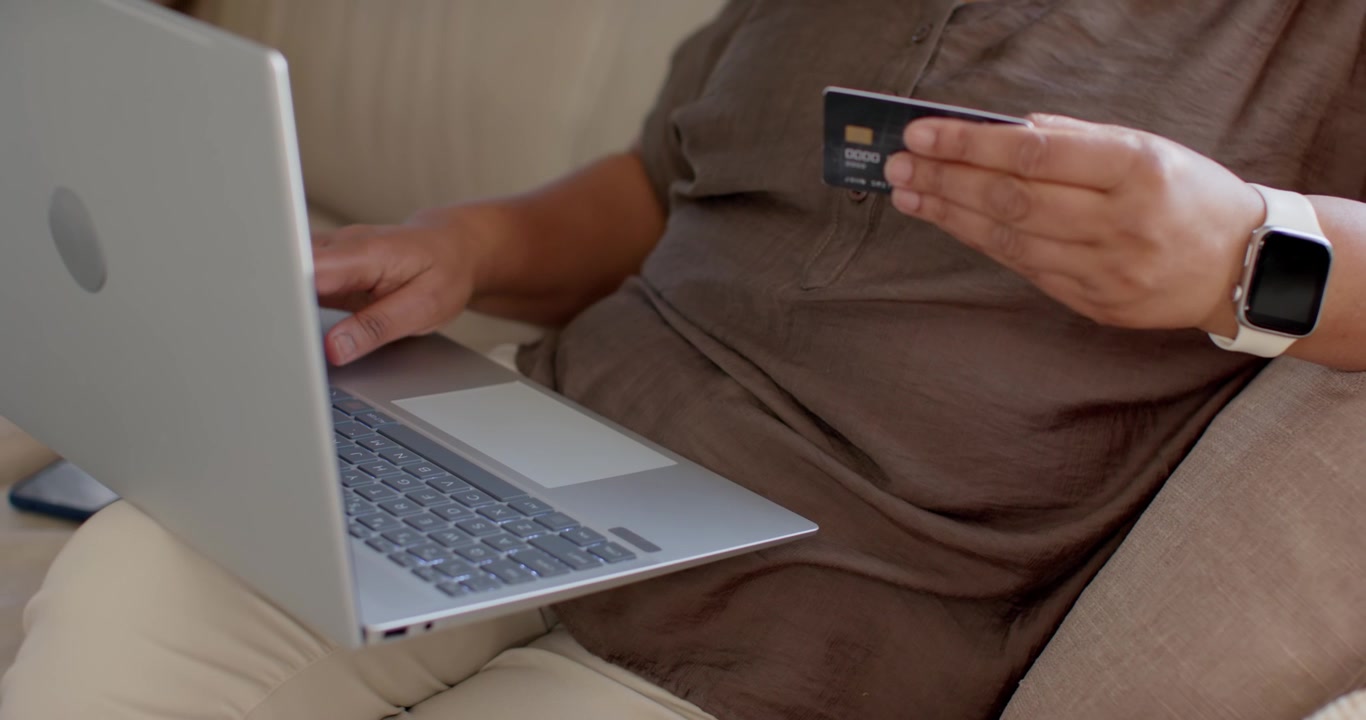 Un adulto inserisce i dettagli di pagamento sul laptop dopo aver controllato il chip della carta, mentre è seduto su un divano beige.