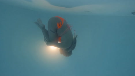 cinematic shot of a woman diving in a fetal position while wearing an orange bikini and vintage diving goggles filmed agains an underwater light close to the water surface in 4k, 120 fps, slomo