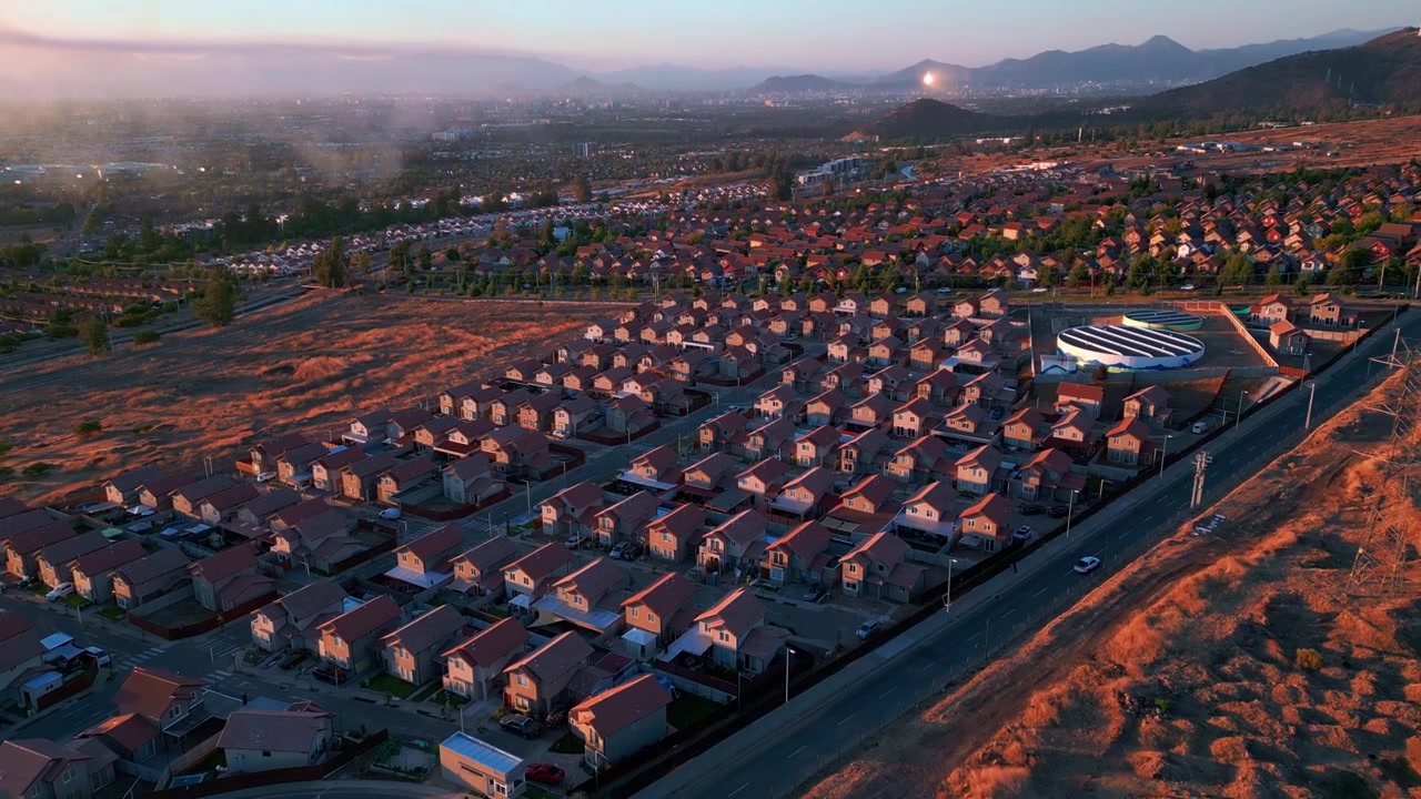 Tilt down aerial shot over dense uniform residential suburb surrounded by dry arid hillsides and misty valley with mountain foothills glowing in warm golden hour light