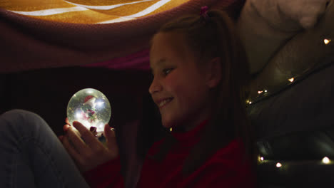 caucasian girl smiling while holding snow globe under blanket fort during christmas at home