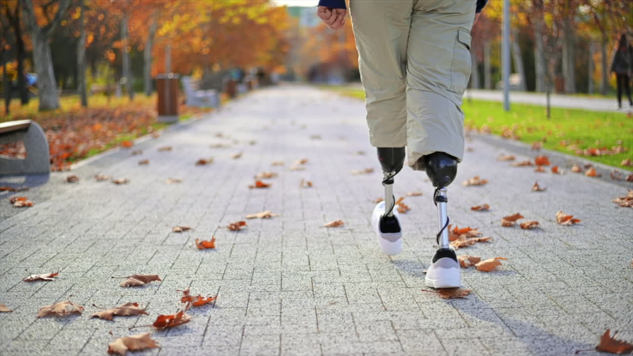 Slow motion view of a walking man with prosthetic legs and white sneakers in a park. Fallen yellow leaves on the ground