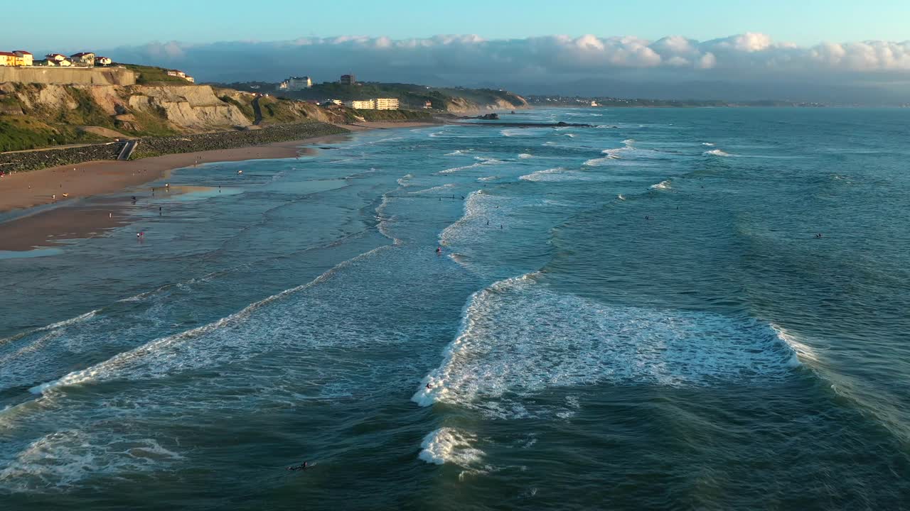 Scenic aerial drone shot of Biarritz coastline, with surfers in Atlantic waves, sandy beach, and cliffs under partly cloudy sky. Popular French surf spot, France